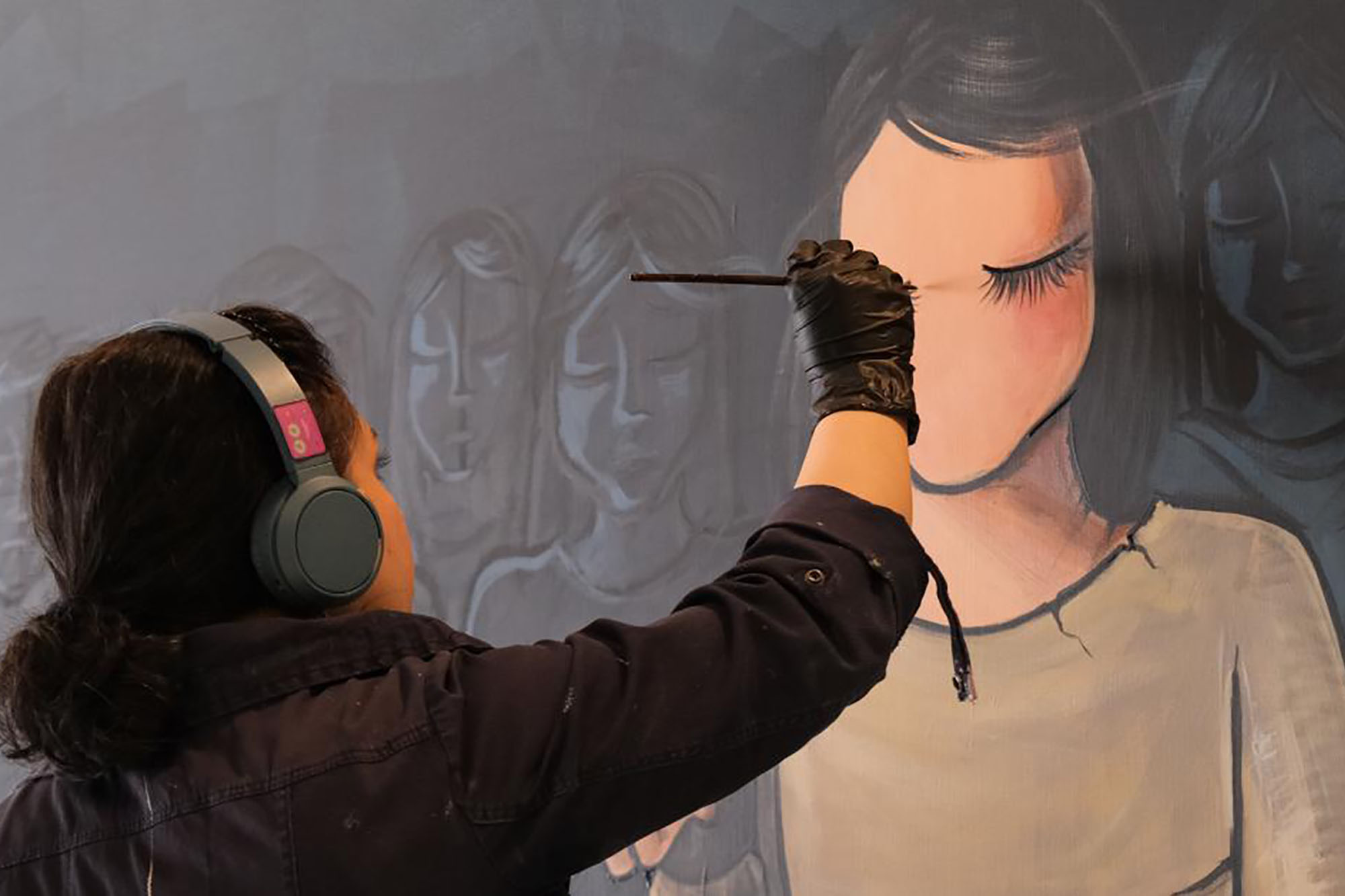 A woman wearing headphones and painting a mural depicting a girl.